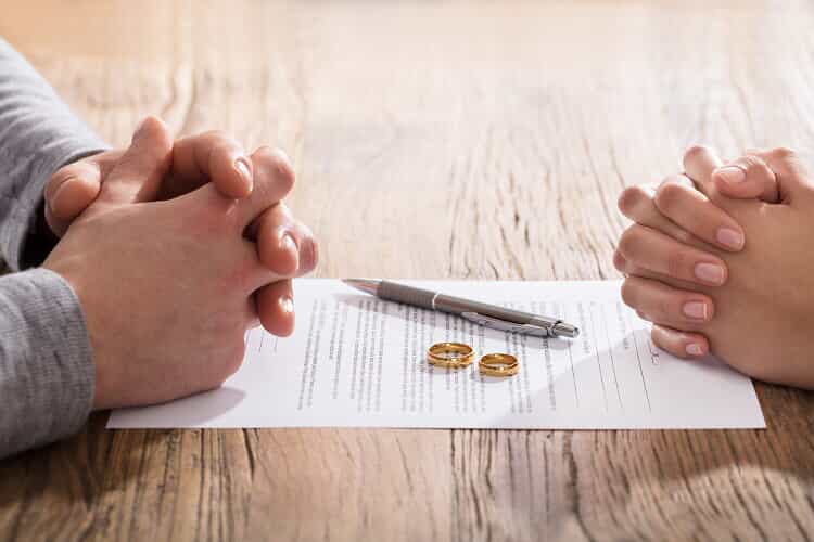 The folded hands of a husband and wife sit on top of DIY divorce papers with their gold wedding rings and a pen